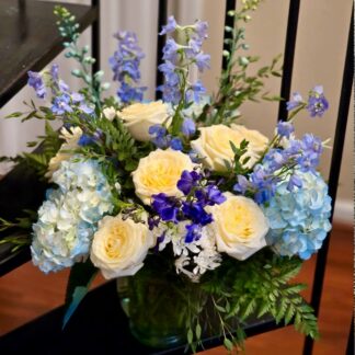 A bouquet in a glass vase with a white fragrant rose, blue hydrangea, blue delphinium, spray white chrysanthemum, and fern