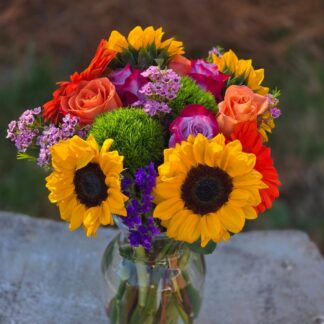 A bouquet in a glass vase with sunflowers, roses, dianthus, chamelacium, statice, and orange gerbera is a sunny joy for any occasion