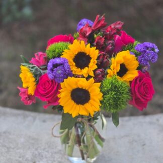 A bouquet in a glass vase with sunflowers, green dianthus, red alstroemeria, pink roses and Monte Casino