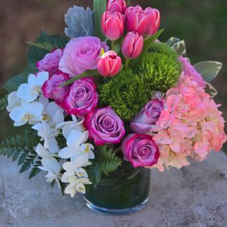 A bouquet in a glass vase with pink hydrangea, green dianthus, lilac rose, orchid, and pink tulips