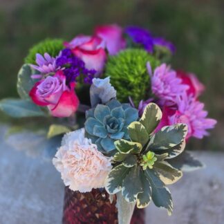 A bouquet in a glass colored vase with a lilac rose, green dianthus, pink carnation, spray pink chrysanthemum, and statice